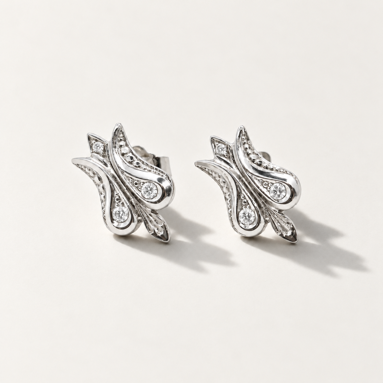 A pair of white gold stud earrings with an intricate, stylized paisley or leaf-like design set with small diamonds, photographed on a clean, light beige background.